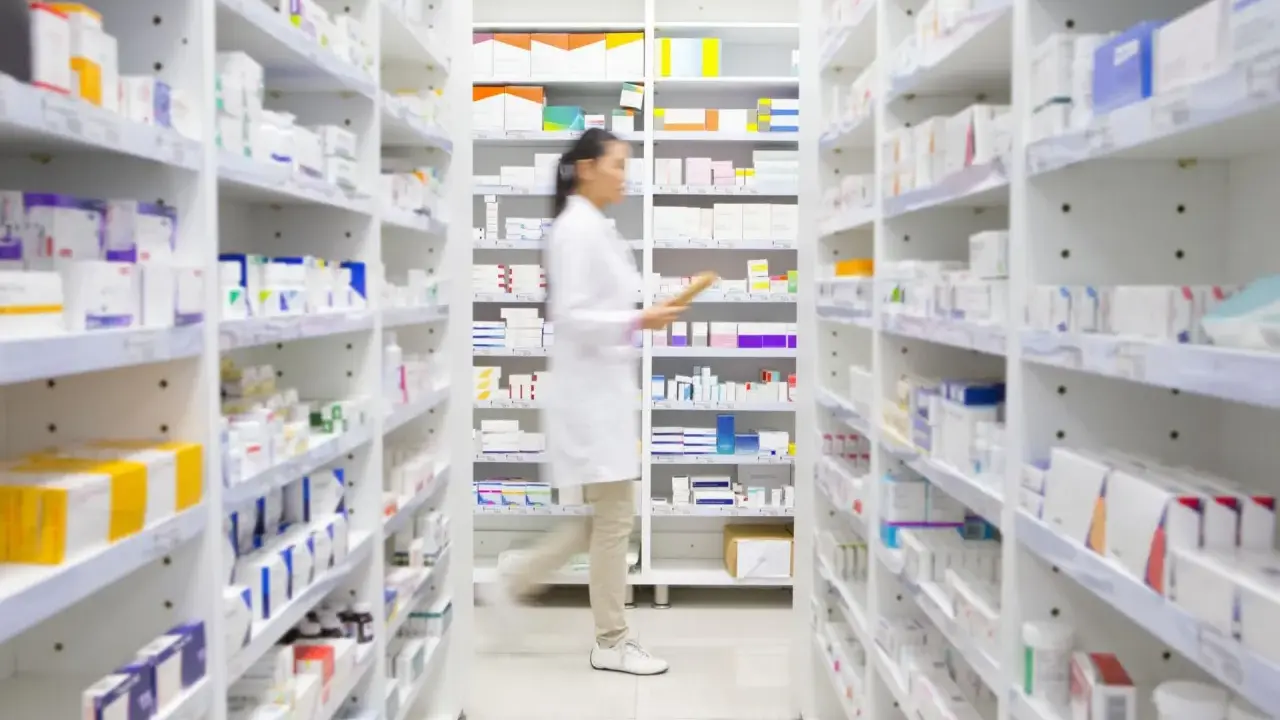 Farmaceutka w białym fartuchu idzie między regałami pełnymi leków. Widać, na co **emanera na co** się przygotowuje, zapewne do wydania pacjentowi.