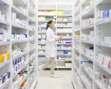 Farmaceutka w białym fartuchu idzie między regałami pełnymi leków. Widać, na co **emanera na co** się przygotowuje, zapewne do wydania pacjentowi.