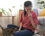 Dziewczyna zatyka nos, czując objawy alergii na kota. Kot siedzi obok niej na dywanie.